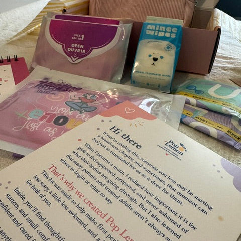 A curated first-period kit laid out on a bed, including a pink corduroy bag, a blue box of wipes, folded paper goods, and a printed card addressed 'Hi there' with text and a heart.