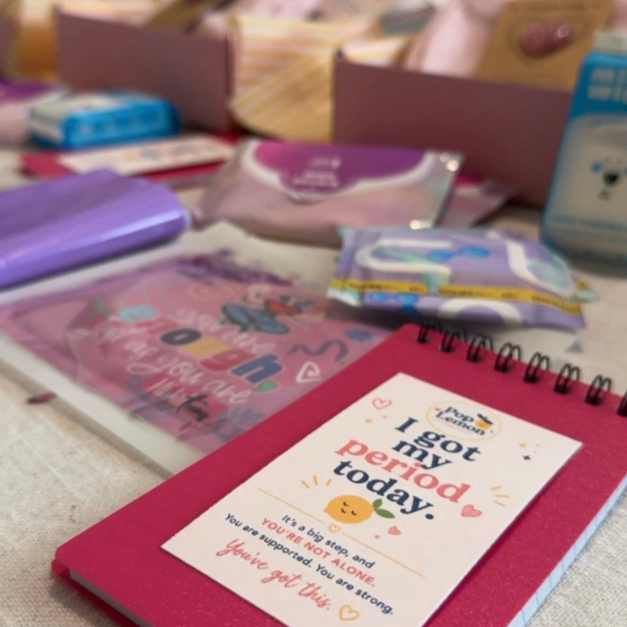 A pink spiral notebook with a white label that reads 'I got my period today.' in colorful text, placed on a table surrounded by other pastel-colored stationery items and boxes.